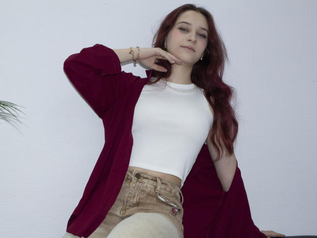 TorriAustin with red hair, white top, and red open shirt, wearing beige shorts, hand near chin, looking slightly down, soft smile, pale skin, subtle makeup, small earrings, bracelet.