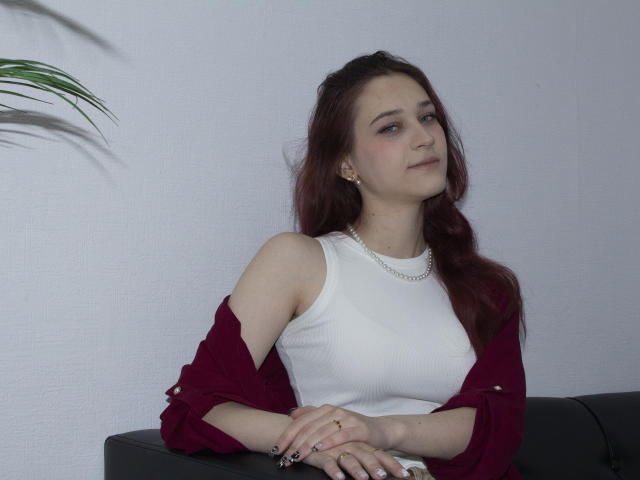 TorriAustin with red hair, white tank top, pearl necklace, and maroon shirt, sitting on black couch, hands crossed, slight smile, white wall, green plant in background.