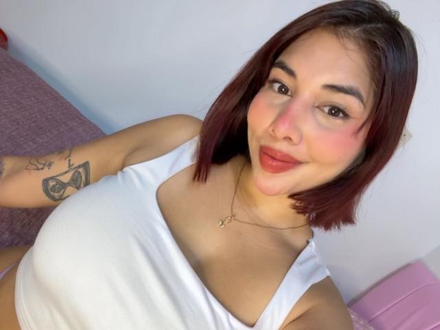 LuciMarteli has red-tinted brown bob, white tank top, showing tits, tattoo on right arm, light skin, subtle smile, pink lipstick, necklace with small cross, pink bed in background.