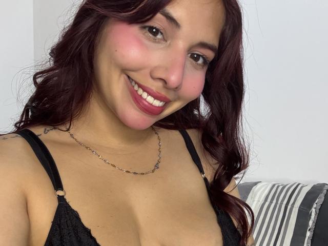 LuciMarteli with red wavy hair, pink blush, smiling, wearing black lace bra, silver necklace, and pink lipstick, showing her tits and cute face, looking at the camera.
