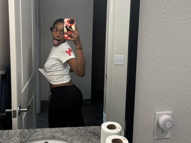 Ouyaya's ass is in black pants, she's sticking her tongue out, wearing a white shirt with a red cross, holding a phone with a red and white case.