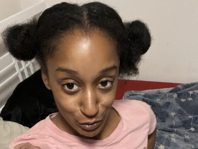 Ouyaya with dark skin, puffy black double buns, and a goofy expression, wearing a pink shirt, gazing up at the camera with slightly parted, shiny lips.