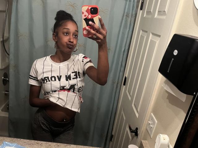 Ouyaya with dark skin, small breasts, and a flat stomach, wearing a white "Slow New York" crop top and gray pants, taking a mirror selfie in a bathroom.