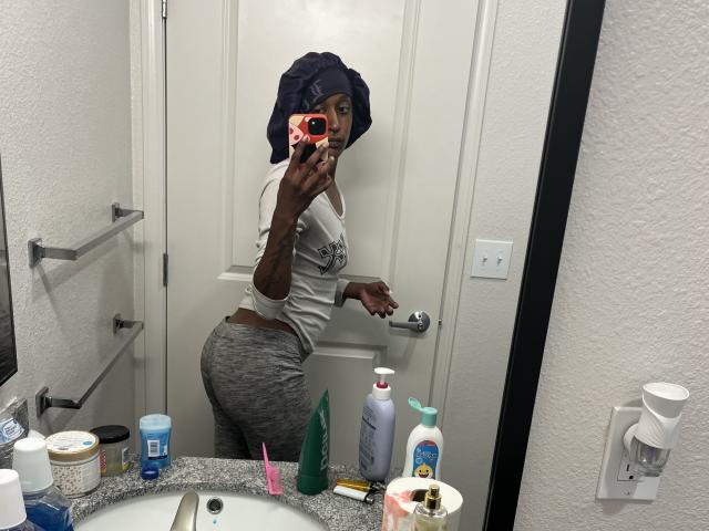 Ouyaya with dark skin, curly hair in a towel, white tee, gray pants, showing her ass in a bathroom mirror selfie. Bathroom sink with various toiletries in the foreground.