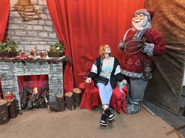 SarahRousen with Santa, sitting on red chair, Santa holding bottle, SarahRousen looking up, Santa's big ass, red curtain, brick fireplace, wood logs, Christmas decorations, black sneakers.