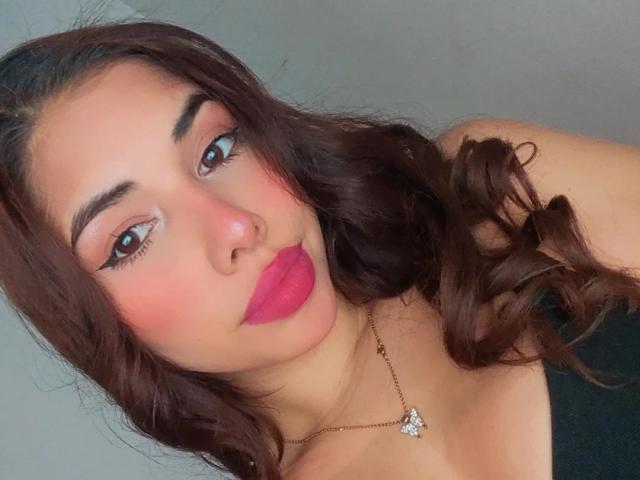 LuciMarteli has pink lipstick, brown curly hair, and a necklace with a diamond. She's wearing a black top, and her tits are slightly visible. She's looking at the camera.