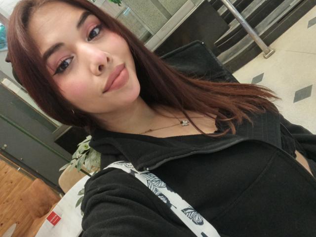 LuciMarteli with pouty lips, brown hair, pink eyeshadow, black top, black hoodie, necklace, white and black bag, sitting in a room, looking at camera.