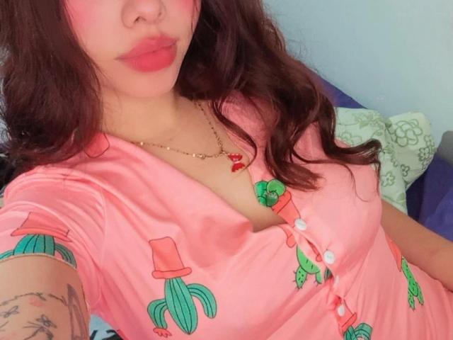 LuciMarteli with pink dress, cactus prints, buttons, and cleavage, red lipstick, wavy brown hair, tattoo on arm, gold necklace, pouty lips, and cute face, sitting on blue bed.