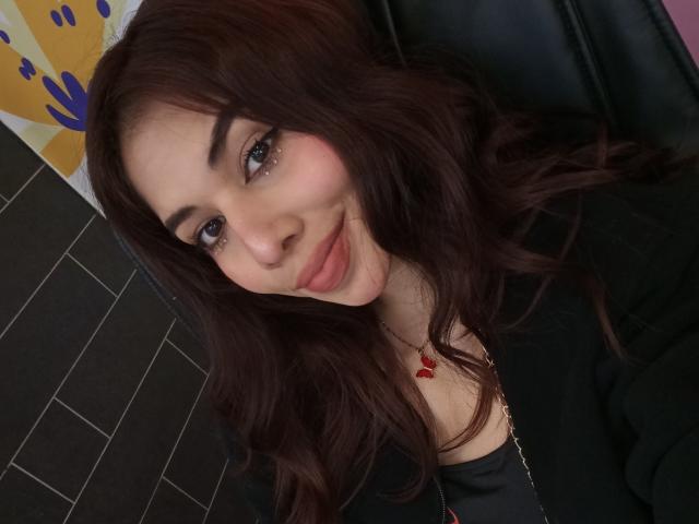 LuciMarteli with wavy dark brown hair, light skin, and full lips, smiling softly at the camera, wearing a black top, and a red heart necklace.