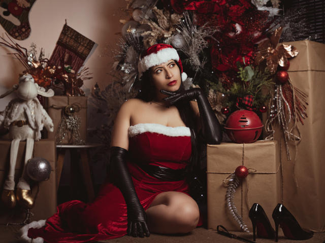 KamileNilsen with red velvet Santa dress, black gloves, and Santa hat, sitting with hand on cheek, showing her tits, surrounded by Christmas decor, gifts, and black heels.