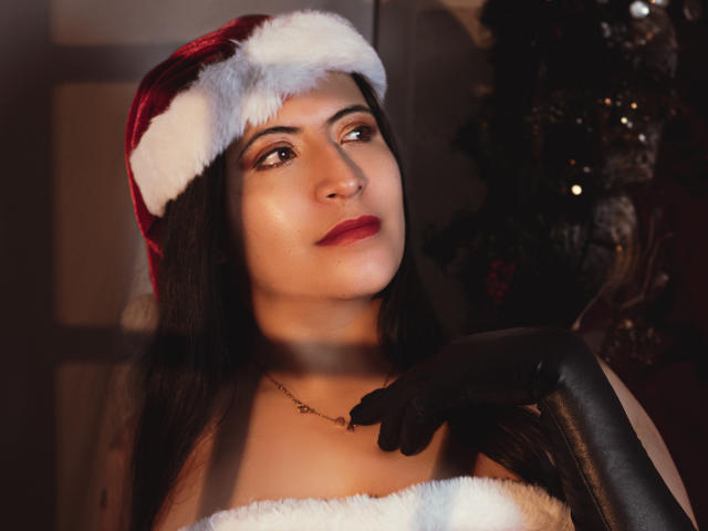 KamileNilsen with dark hair, wearing a red Santa hat with white fur, black gloves, and a white fur strap dress, looking up with red lipstick, and a slight smile.