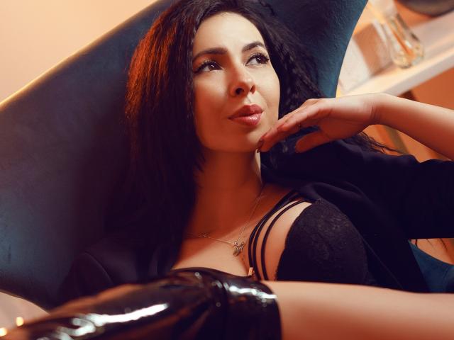 ChloeStone with long black hair, wearing a blue denim off-shoulder shirt, looking over her shoulder with a slightly pouty expression, hands near her neck.