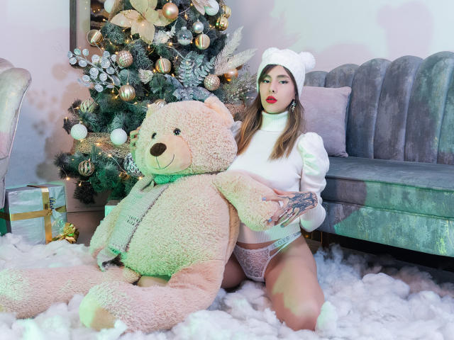 NathalyJohns mit nacktem Hintern, die einen weißen Hut und einen weißen Pullover trägt, sitzt auf einem flauschigen weißen Teppich, hält ein Geschenk und befindet sich neben einem riesigen Teddybären, in der Nähe eines geschmückten Weihnachtsbaums..