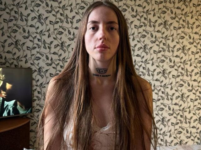 AdrianaCult has long brown hair, a tattoo under her chin, and is wearing a white lace top, showing her tits, with a serious expression in a room.