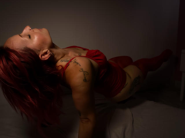 GiselleDunn with red hair, red lingerie, and tattoos, lying back with eyes closed, head tilted, arms above, legs bent, in dim lighting, showing her ass and tits.