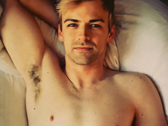 Shirtless PartyTips with blonde hair, blue eyes, and a slight smile, lying on a bed with an arm raised, showing a hairy armpit. His chest is bare.