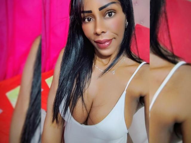 PamelaSweetCoquine has long black hair and brown skin. She's wearing a white, low-cut tank top that reveals significant cleavage. She's looking at a mirror, with her reflection visible on the right. She has a slight smile, dark eyebrows, and wears a small necklace. The background is pink and red. The image is a photograph.