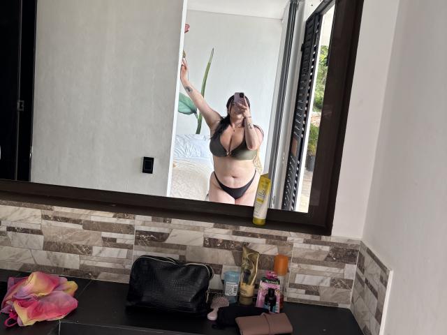 VenusxMusa with big tits in a green bra and black panties, standing in front of a mirror, holding a phone, in a bathroom with a dark wooden frame.