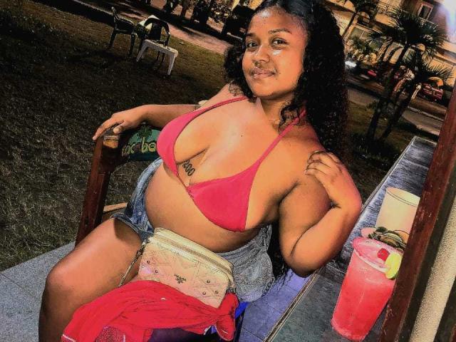 OshayaDivine, a plus-sized woman with curly black hair, sits in a wooden chair at a night-time outdoor event. She wears a pink bikini top that reveals her large breasts and a tattoo under her left breast. She has a slight smile on her face. A red cloth is draped over her lap, and a pink drink is on a table to her right. The background is blurred, focusing on her. The image is brightly lit, emphasizing her brown skin and the vibrant pink of her bikini top.
