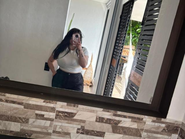 VenusxMusa, avec de longs cheveux noirs, une chemise blanche, un pantalon noir taille haute, tenant un téléphone, debout devant un miroir, montrant ses fesses, dans une salle de bain avec un mur en carrelage..
