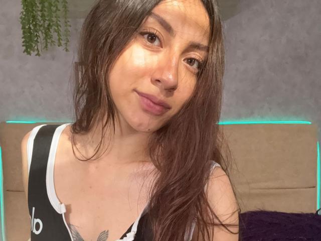 Luluberrie has long brown hair, a slight smile, and a tattoo on her chest. She's wearing a black and white top with straps, showing her small tits.