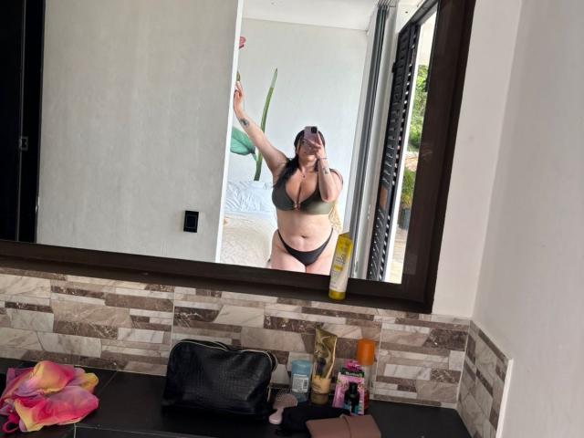 VenusxMusa with big tits and black panties, wearing a green bra, taking a mirror selfie, holding a pink flower, standing in a bathroom with a black bag.