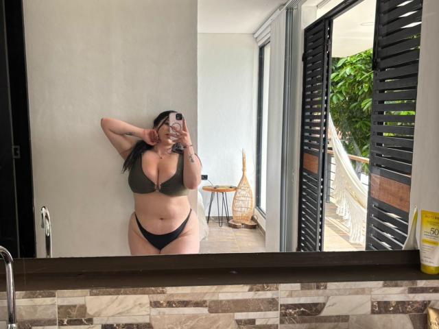 VenusxMusa with big boobs, wearing an olive green bra and black thong, standing in a bathroom, taking a mirror selfie, her right hand in her hair.