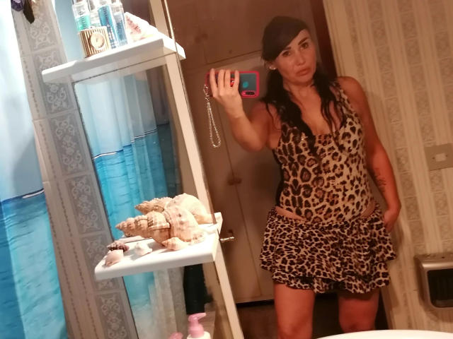 VictoriaFoxx-hot with long dark hair, wearing a leopard print dress, standing in a bathroom, holding a pink phone, mirror selfie, showing a small, flirty ass, and a cute face.