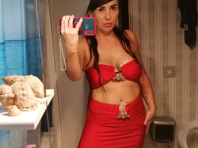 VictoriaFoxx, sensual em um top vermelho sem alças e uma saia combinando, exibindo seus seios e um piercing no umbigo, tira uma selfie no espelho com um celular rosa..