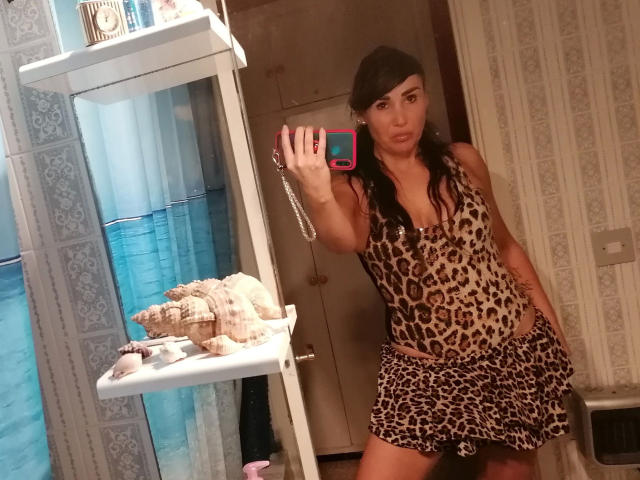 VictoriaFoxx – attraktiv in einem Leopardenmuster-Kleid, stehend in einem Badezimmer, mit einem rosa Telefon in der Hand, mit langen, dunklen Haaren und einem selbstbewussten, leicht schmollenden Ausdruck, wobei sie ihr Hinterteil zur Schau stellt..