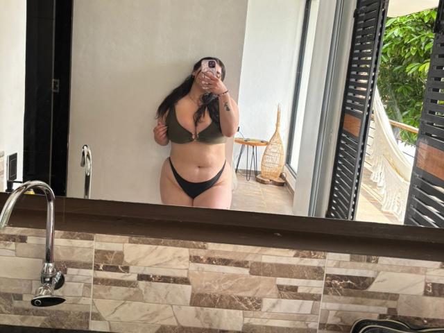 VenusxMusa with large boobs and a curvy ass, wearing an olive green bra and black thong, taking a mirror selfie in a bathroom with tiled walls.