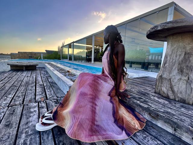 KharlaQueen, con piel oscura y trenzas largas, vestida con un vestido de color rosa y morado con estampado tie-dye, sentada en una terraza de madera, con chanclas blancas, y una casa de vidrio moderna al fondo..