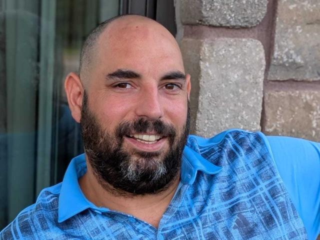 Erazion, bald with a full beard, wearing a blue plaid shirt, smiling slightly with visible teeth, standing in front of a stone wall.