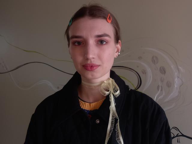 LisaDarcy with pale skin, brown hair in a bun, red and orange hair clips, wearing a black jacket, choker, and yellow top, looking at the camera with pink lips.