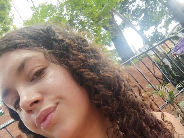 Close-up of RubbyStrongX with curly brown hair, light brown skin, and subtle makeup. RubbyStrongX has a slight smile, full lips, and visible freckles. The background includes green leaves, a metal fence, and blurred outdoor elements. The image has natural light, emphasizing the texture of RubbyStrongX's hair and skin. The composition focuses on RubbyStrongX's face, with the background slightly out of focus. RubbyStrongX's relaxed expression and casual setting are prominent.