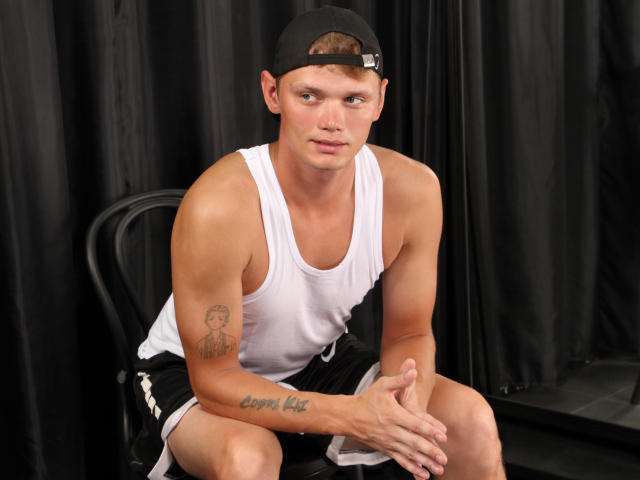 BillbateMan, muscular, white tank top, black cap, black shorts, tattoo on right arm, tattoo on left forearm, black curtain background, sitting on black chair, hands on knees.