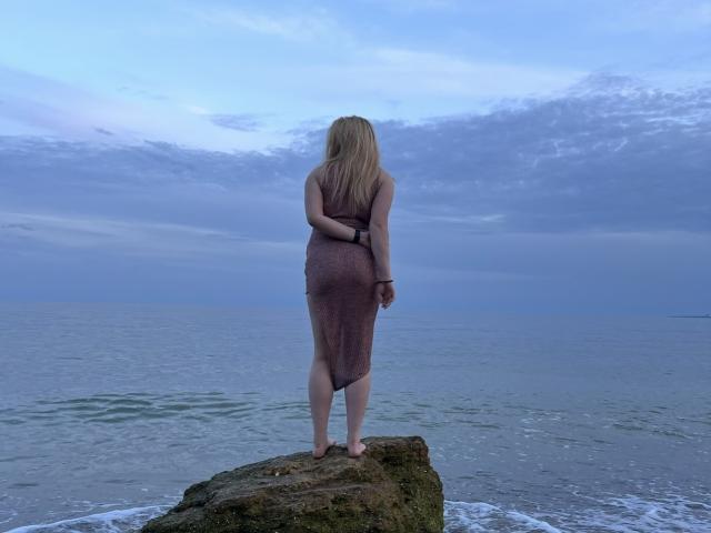 Alice Smity, avec ses longs cheveux noirs, un haut blanc remonté, exposant ses seins nus et ses mamelons, assise sur un lit avec une tête de lit vert turquoise et des oreillers noirs..