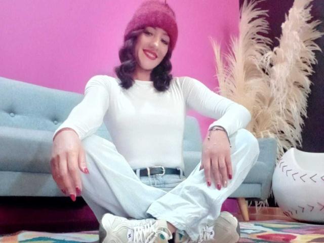 Mulher com cabelo loiro, camisa rosa e jeans azuis com apliques de flores, mostrando o bumbum em uma selfie no espelho, em pé ao lado de um sofá bege e uma bola de exercícios..