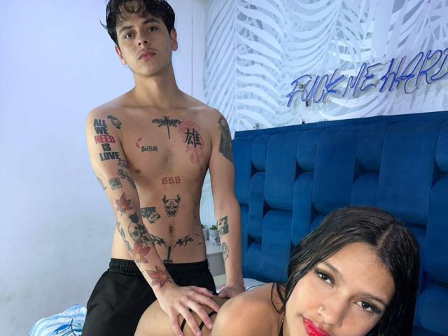 SamanthaCorine sitzt mit angelegten Beinen, wobei ihre weißen Brüste durch ein enges Hemd sichtbar sind. Sie trägt eine rosa Mütze, helle Jeans und weiße Turnschuhe und lächelt mit roten Nägeln..