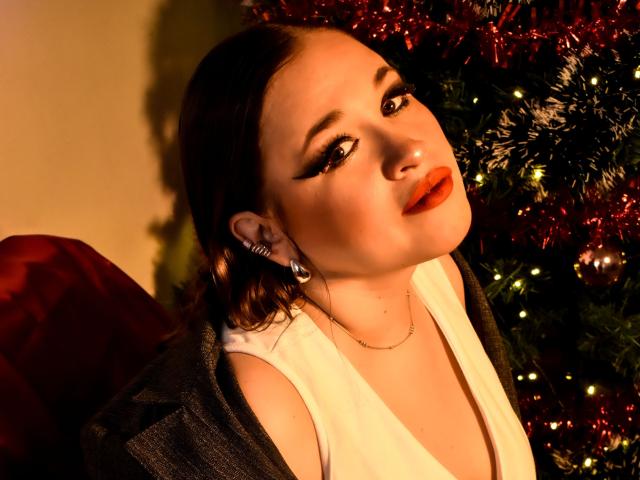 MillyGrey con lápiz labial rojo, ojos cerrados, manos en un blazer marrón, chaleco blanco, aretes de perlas, de pie frente a un árbol de Navidad, con los brazos levantados, luciendo sexy, mostrando sus senos..