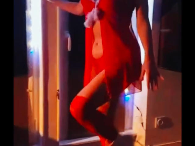 MillyGrey with red scarf, white sleeveless dress, tits showing, head tilted back, lips parted, looking up, holding scarf, Christmas tree with lights and ornaments in background.