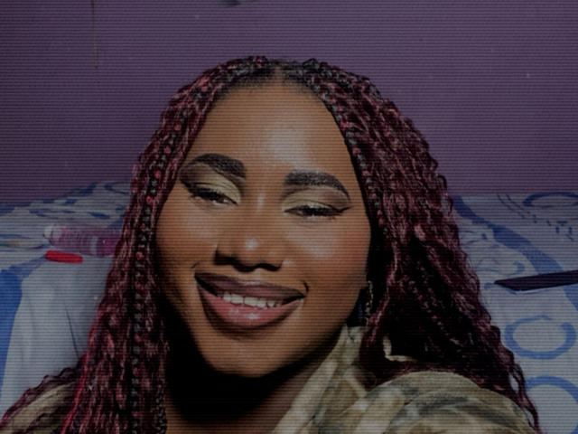 Dilay hat eine dunkelbraune Haut, rote Zöpfe und lächelt mit halb geschlossenen Augen. Sie trägt Make-up und ein grünliches Hemd und sieht süß und attraktiv aus..