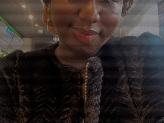 Dilay's face with a slight smile, wearing a dark, textured, zip-up jacket. She has short curly hair, brown skin, and is looking slightly to the side.