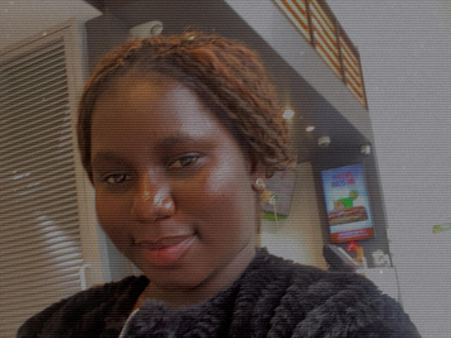 Dilay with dark skin, brown braided hair, small nose, smiling slightly, wearing a black textured shirt, small earrings, in a dimly lit room.