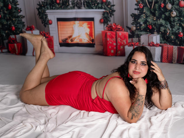 KathyFlores, curvy with large ass, lying on white bed in red dress, legs up, tattooed arm, black curly hair, red lipstick, fire in background, Christmas trees, presents.