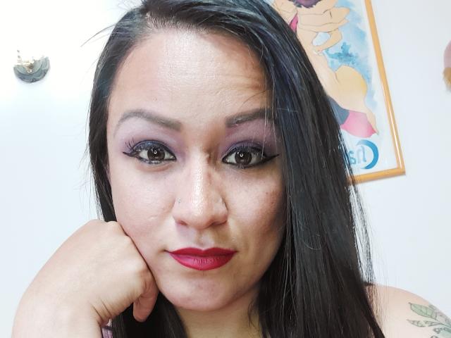 LucianaDia with black hair, purple eyeshadow, red lipstick, and black eyeliner, resting her chin on her hand, showing a tattoo on her left shoulder. Her tits are small.