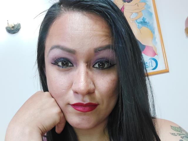 LucianaDia with black hair, purple eyeshadow, red lipstick, and black eyeliner, resting her chin on her hand, showing tattoos on her arm, looking at the camera.
