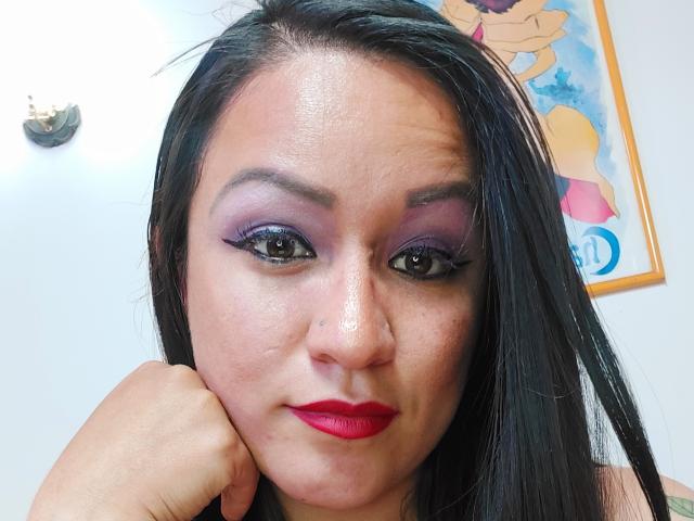 LucianaDia has dark straight hair, purple eyeshadow, black eyeliner, red lipstick, and a small nose piercing. Her hand rests on her cheek, showing a slight smirk.