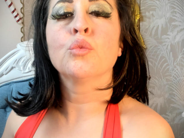 ScarletMilfX, con cabello negro, una blusa roja y ajustada, y sombra de ojos verde brillante, frunce los labios en un selfie. Sus pechos son parcialmente visibles. Ella está guiñando un ojo..
