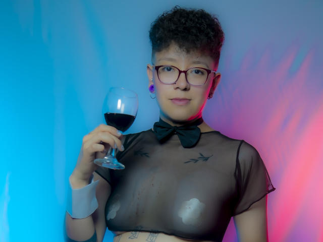 AllexNoa met krullend haar, een bril, en een zwarte doorschijnende top die haar borsten laat zien, houdt een glas rode wijn vast, met blauw en roze licht op haar..
